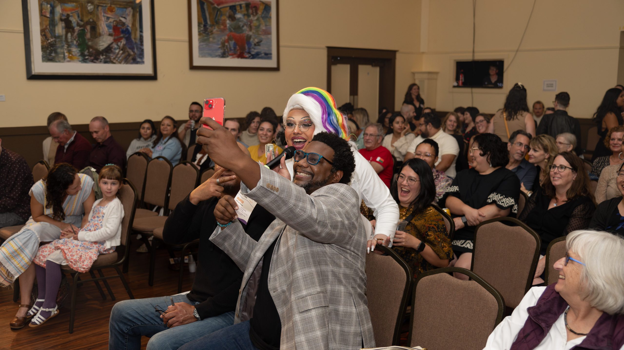 A smiling Black man is posing with a drag queen in a rainbow wig for a selfie, with the audience smiling behind them.